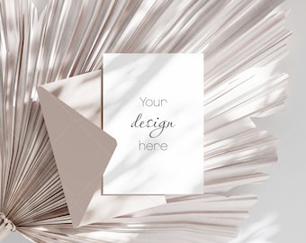 Puede incluir: Una tarjeta blanca en blanco con el texto "Your design here" descansa sobre un sobre beige, sobre un fondo de una hoja de palma seca. La escena está bañada en luz suave, creando sombras.