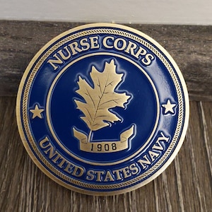 Collectible military challenge coin that has a blue background, oak leaf in the middle with a banner stating 1908, and Nurse Corps United States Navy written in antique gold around the outline.