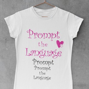 Puede incluir: Camiseta blanca con el texto "Prompt the Language" en letras rosa y negras. Un corazón rosa con una pieza de rompecabezas dentro está por encima del texto.