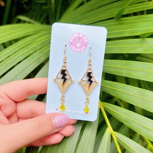 May include: Gold-tone earrings with a geometric design featuring a black lightning bolt. The earrings have yellow bead accents and are displayed on a light blue card with the text "ROSE & CO JEWELRY". The background is lush green foliage.