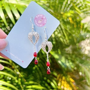 May include: Silver-coloured angel wing earrings with three red teardrop-shaped beads dangling from each earring. The earrings are displayed on a light blue card with a pink logo that says "Rose Jewelry Co." and the words "Handmade with Love."