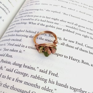 Puede incluir: Un anillo artesanal de alambre de cobre con tres piedras de unakita verdes y rosas. El anillo está sobre un libro abierto con texto impreso. El diseño del anillo presenta una banda de alambre enrollado.