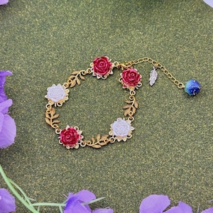 May include: Gold-toned bracelet featuring red and white rose-shaped charms. The bracelet has a delicate chain with leaf accents and a blue and gold bead charm. The bracelet is set against a green background with purple floral accents.