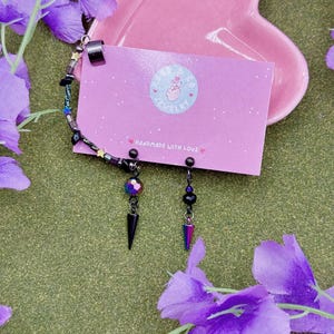 May include: A pair of black earrings with a spike and a colorful, iridescent bead. The earrings are displayed on a pink card that reads "HANDMADE WITH LOVE". A bracelet with black beads and a silver cuff is also visible.