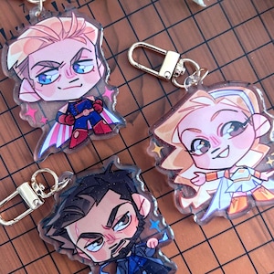 Könnte beinhalten: Drei durchsichtige Acryl-Schlüsselanhänger mit Chibi-Stil Charakterillustrationen. Einer zeigt eine blonde Figur in einem blau-roten Anzug mit Umhang, ein anderer eine blonde Figur in einem weiß-goldenen Outfit und der dritte eine dunkelhaarige Figur in einem blauen Mantel.