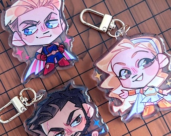 May include: Three clear acrylic keychains featuring chibi-style character illustrations. One depicts a blonde figure in a blue and red suit with a cape, another a blonde figure in a white and gold outfit, and the third a dark-haired figure in a blue coat.