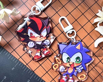 Op de afbeelding: Twee acryl sleutelhangers met stripfiguren. Eén sleutelhanger toont een zwart en rood personage met een boze uitdrukking. De andere sleutelhanger toont een blauw personage met een vrolijke uitdrukking en gouden ringen. Beide sleutelhangers hebben zilveren sluitingen.