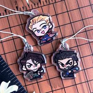Puede incluir: Tres colgantes de acrílico transparente con ilustraciones de personajes estilo chibi. Cada colgante incluye un cordón blanco para colgar. El colgante superior muestra un personaje rubio, mientras que los dos inferiores son morenos.