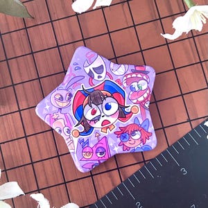以下が含まれることがあります： 星型の紫色のボタンで、漫画のキャラクターのイラストが描かれています。 ボタンは光沢のある仕上げで、紫、赤、青、白の色合いのさまざまな顔が特徴です。 ボタンは茶色の表面にあります。