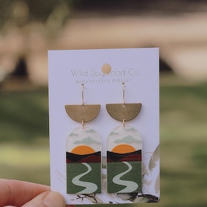 May include: A pair of handmade earrings featuring a sunset landscape with a winding road and a gold crescent moon charm. The earrings are on a white card with the text "Wild Dogwood Co. Handcrafted Jewelry".