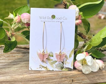 May include: A pair of gold hoop earrings with pink butterfly charms. The earrings are displayed on a white card with the text "Wild Dogwood Co. Handcrafted Jewellery".