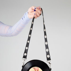 Puede incluir: Un bolso de vinilo negro en forma de disco con una correa de cadena plateada y una correa de cuero negro con ojales plateados. El disco tiene el título "Can't Keep A Good Man Down" de Bee Gees.