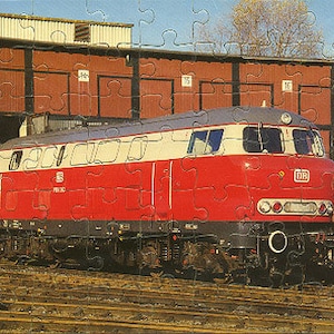 Puede incluir: Una locomotora diésel roja y blanca con las letras "DB" en el lateral. La locomotora está estacionada frente a un edificio de ladrillo con varias puertas de garaje.