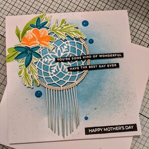 May include: A white card with a blue and white dreamcatcher with a floral design in the top left corner. The card has the text "YOU'RE SOME KIND OF WONDERFUL" and "HAVE THE BEST DAY EVER" on it. The card also has the text "HAPPY MOTHER'S DAY" on it. The card is on a pink surface with the text "hugs enclosed" on it.