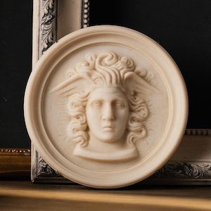 May include: A circular off-white relief plaque featuring a sculpted human face with winged hair. The central figure displays detailed facial features and flowing locks of hair with small wings extending from the sides, all set within a raised border.