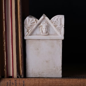 Könnte beinhalten: Eine weiße, rechteckige Marmorbox mit einem dreieckigen Deckel, der ein geschnitztes Gesicht und florale Designs aufweist. Die Box steht auf einem Regal mit antiken Büchern und erzeugt eine klassische Ästhetik.