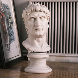 May include: A white marble bust of a Roman figure, featuring detailed facial features and short, curly hair. The bust is mounted on a pedestal with the inscription "C. SVLLA". The sculpture is placed on a wooden floor, with framed artwork and a radiator in the background.