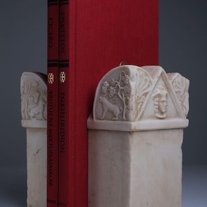 Puede incluir: Dos libros de tapa dura rojos titulados Cicero Brutus ile Mektuplaşmalar y Epiktetos Enkheiridion se sostienen entre dos sujetalibros de piedra de color blanco roto. Los sujetalibros presentan relieves tallados de un lobo, vides y un rostro humano.