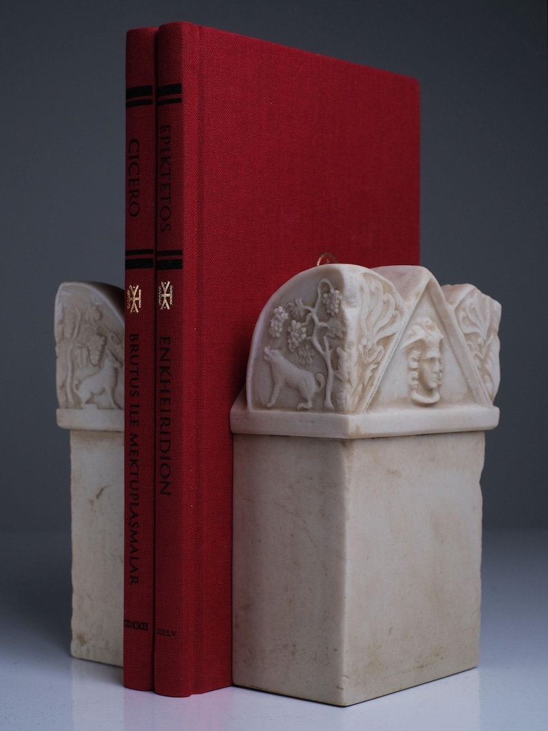 Puede incluir: Dos libros de tapa dura rojos titulados Cicero Brutus ile Mektuplaşmalar y Epiktetos Enkheiridion se sostienen entre dos sujetalibros de piedra de color blanco roto. Los sujetalibros presentan relieves tallados de un lobo, vides y un rostro humano.