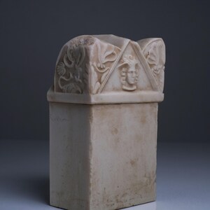 Puede incluir: Una urna funeraria rectangular de piedra color hueso con un front&oacute;n triangular tallado en la tapa. El front&oacute;n muestra un relieve central de un rostro humano flanqueado por intrincadas tallas de volutas florales en los paneles.