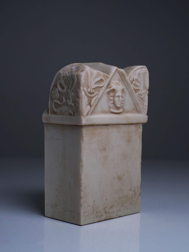 Puede incluir: Una urna funeraria rectangular de piedra color hueso con un front&oacute;n triangular tallado en la tapa. El front&oacute;n muestra un relieve central de un rostro humano flanqueado por intrincadas tallas de volutas florales en los paneles.