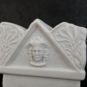 May include: A light grey stone-like sculpture featuring a central relief of a winged head within a triangular pediment. The design includes ornate floral carvings on the flanking sections, all rendered in a matte, monochromatic finish.