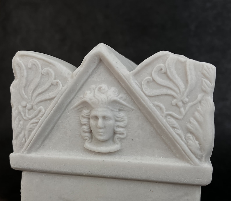 May include: A light grey stone-like sculpture featuring a central relief of a winged head within a triangular pediment. The design includes ornate floral carvings on the flanking sections, all rendered in a matte, monochromatic finish.
