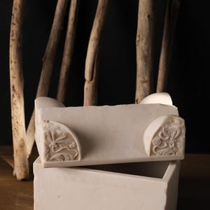 May include: A rectangular white stone box with a removable lid. The lid features carved floral corner details. The box has a smooth, matte finish and a clean, geometric structure, showcasing the natural texture and light colour of the stone material.