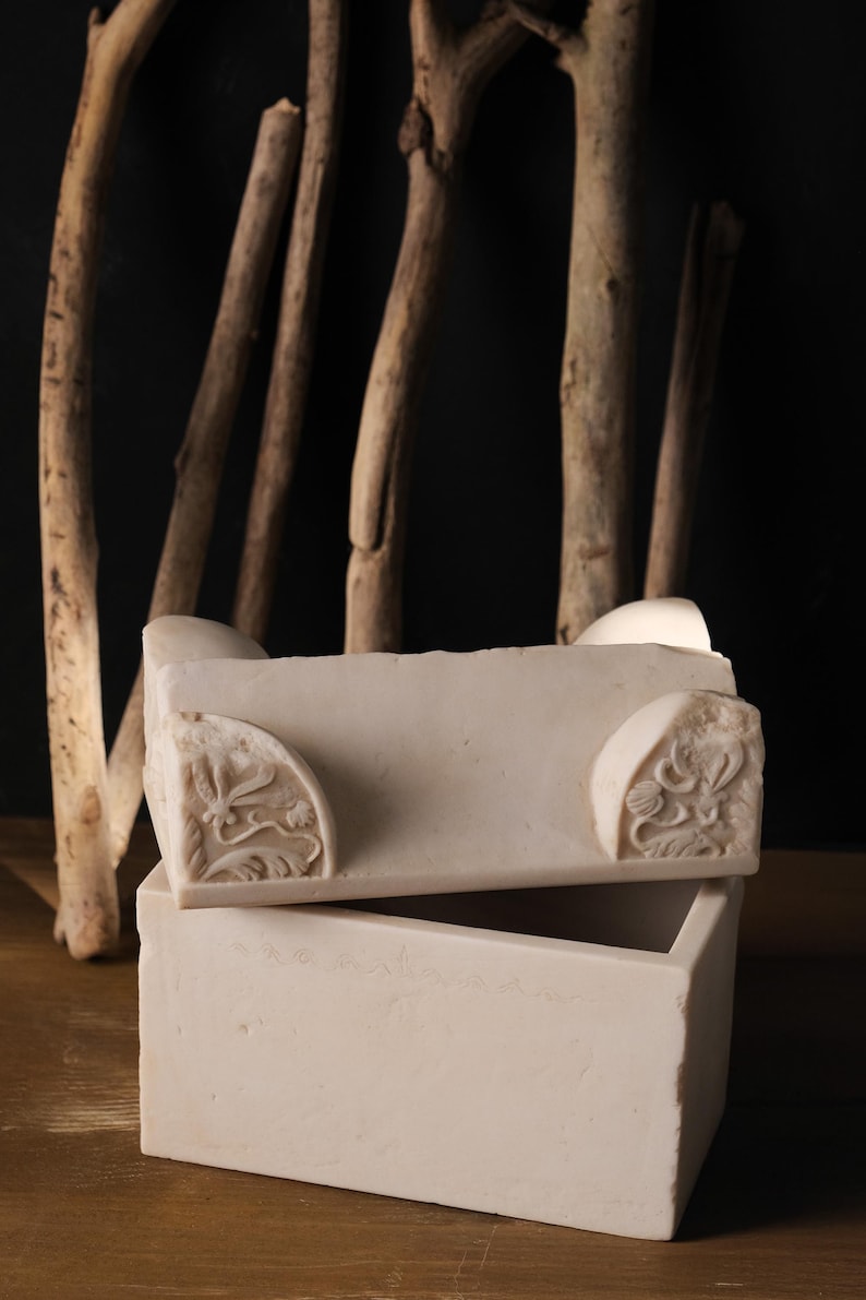 May include: A rectangular white stone box with a removable lid. The lid features carved floral corner details. The box has a smooth, matte finish and a clean, geometric structure, showcasing the natural texture and light colour of the stone material.