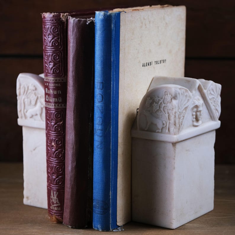 Puede incluir: Dos sujetalibros de m&aacute;rmol blanco con relieves detallados de figuras y follaje, sosteniendo tres libros. Los libros incluyen un volumen rojo oscuro titulado Ruhun &Ouml;l&uuml;m&uuml;, un lomo oscuro liso y un lomo azul con el texto Aleksei Tolstoy.