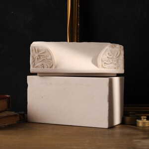 May include: A white stone architectural fragment featuring carved floral and scroll motifs on the sides, resting atop a solid rectangular white stone block. The piece has a smooth, matte finish and displays intricate relief details on both ends.