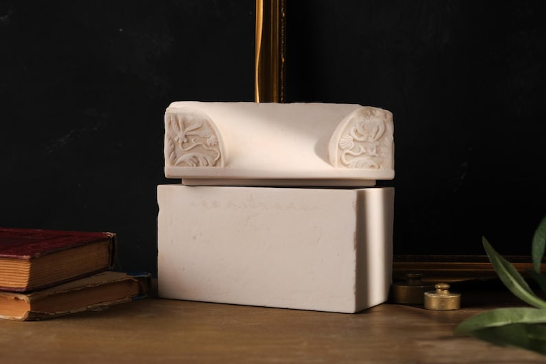 May include: A white stone architectural fragment featuring carved floral and scroll motifs on the sides, resting atop a solid rectangular white stone block. The piece has a smooth, matte finish and displays intricate relief details on both ends.
