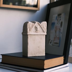 Puede incluir: Una caja rectangular de piedra color hueso con una tapa de front&oacute;n triangular. La tapa est&aacute; decorada con motivos florales tallados en los laterales y un relieve central de un rostro humano con cabello rizado. La base es lisa.