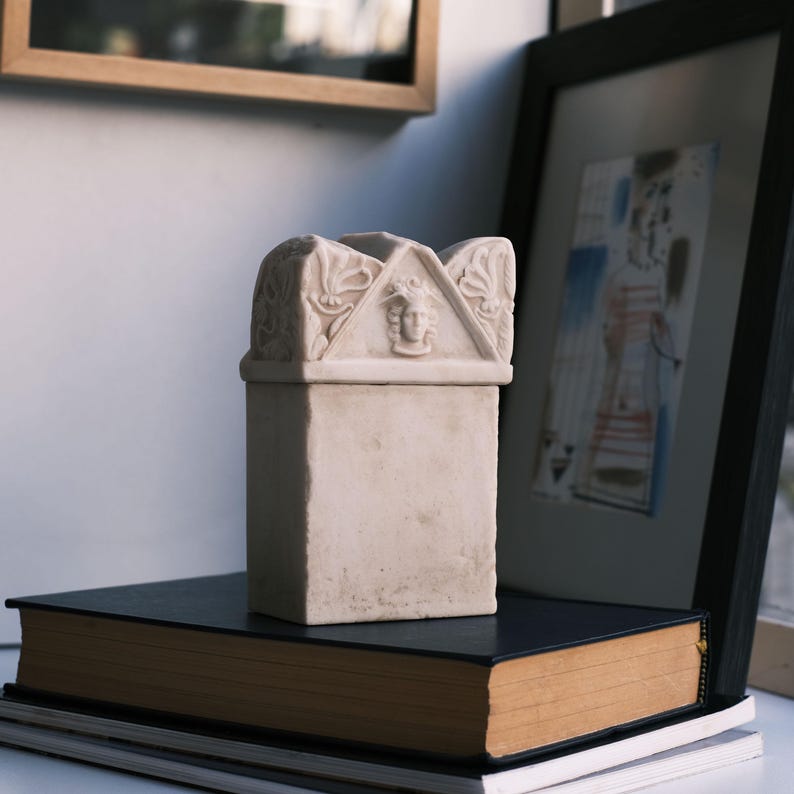 Puede incluir: Una caja rectangular de piedra color hueso con una tapa de front&oacute;n triangular. La tapa est&aacute; decorada con motivos florales tallados en los laterales y un relieve central de un rostro humano con cabello rizado. La base es lisa.