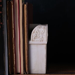 Puede incluir: Un sujetalibros de m&aacute;rmol blanco con un dise&ntilde;o de relieve floral tallado, colocado junto a una fila de libros de tapa dura. Los libros incluyen un volumen titulado The Study of Colour de Jacobs y un libro con la palabra michelangelo.