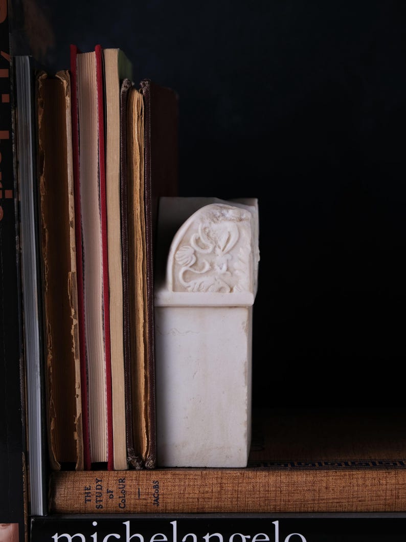 Puede incluir: Un sujetalibros de m&aacute;rmol blanco con un dise&ntilde;o de relieve floral tallado, colocado junto a una fila de libros de tapa dura. Los libros incluyen un volumen titulado The Study of Colour de Jacobs y un libro con la palabra michelangelo.