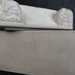 May include: A rectangular off-white stone box with a lid. The lid features carved floral motifs on both ends. The front face of the box displays a horizontal engraved line with a repeating wave-like pattern across the upper portion of the surface.