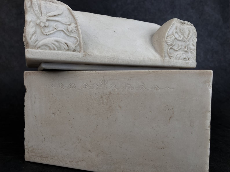 May include: A rectangular off-white stone box with a lid. The lid features carved floral motifs on both ends. The front face of the box displays a horizontal engraved line with a repeating wave-like pattern across the upper portion of the surface.