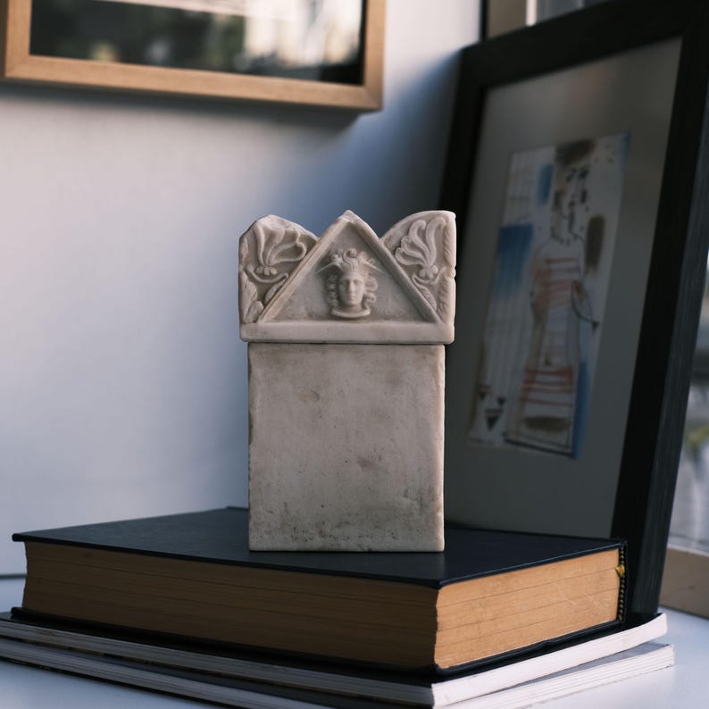 Puede incluir: Un objeto rectangular de piedra color blanco hueso con una base plana y una secci&oacute;n superior decorativa. La parte superior presenta un front&oacute;n triangular con un rostro humano esculpido en el centro y tallas florales laterales, similar a una estela.