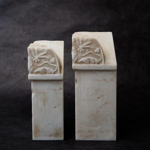 Puede incluir: Dos sujetalibros de piedra color blanco roto con base rectangular y parte superior curva. Cada pieza presenta un patr&oacute;n floral tallado en relieve en el lateral, mostrando hojas y flores delicadas. Las superficies de piedra tienen textura.