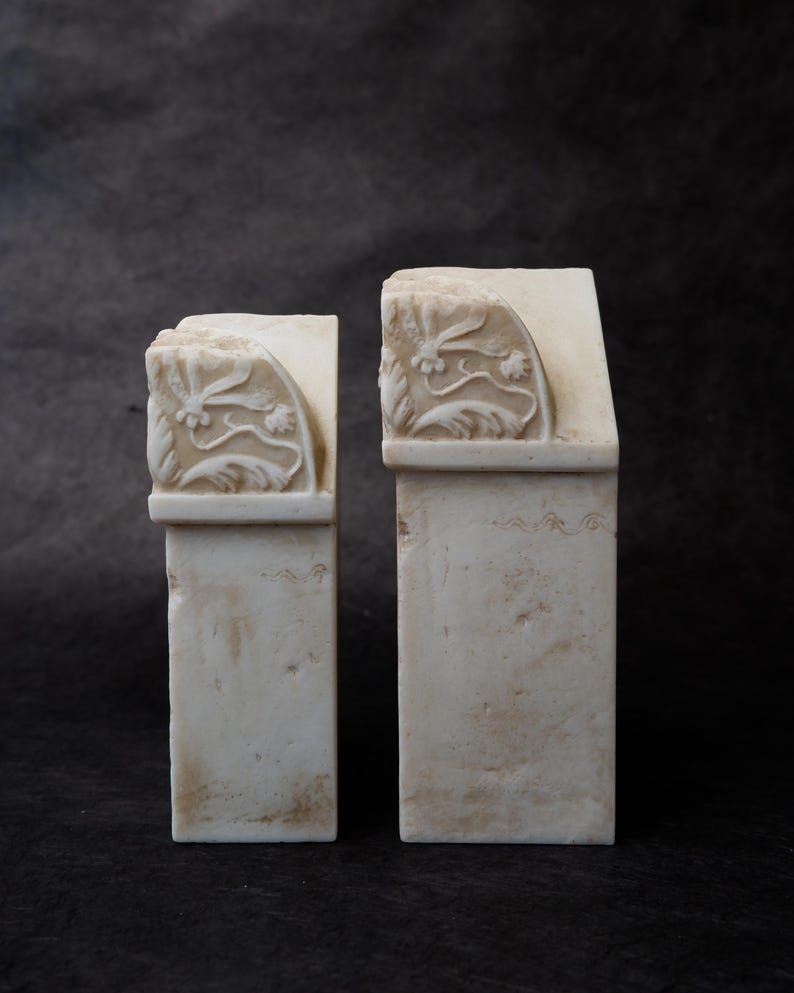 Puede incluir: Dos sujetalibros de piedra color blanco roto con base rectangular y parte superior curva. Cada pieza presenta un patr&oacute;n floral tallado en relieve en el lateral, mostrando hojas y flores delicadas. Las superficies de piedra tienen textura.