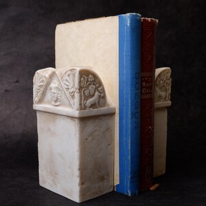 Puede incluir: Un par de sujetalibros de piedra color hueso con detalles en relieve tallados de un rostro humano y un perro entre vides. Los sujetalibros sostienen dos libros, uno con el lomo azul y otro con el lomo de color rojo oscuro.