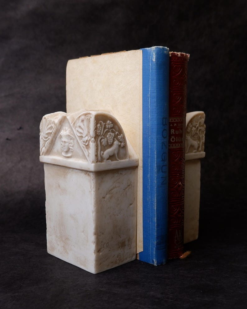 Puede incluir: Un par de sujetalibros de piedra color hueso con detalles en relieve tallados de un rostro humano y un perro entre vides. Los sujetalibros sostienen dos libros, uno con el lomo azul y otro con el lomo de color rojo oscuro.
