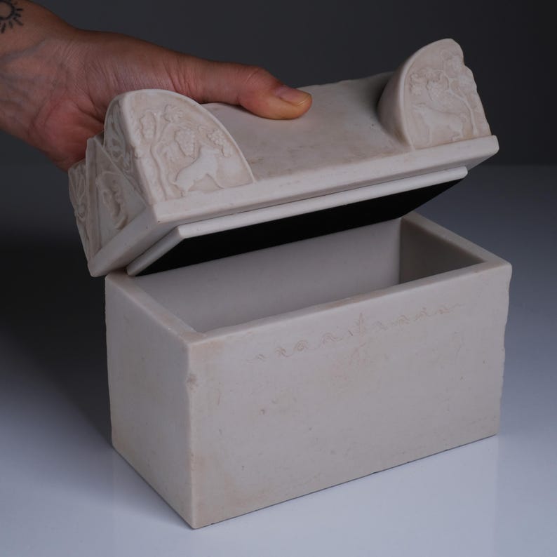 May include: A rectangular off-white stone box with a removable lid. The lid features carved relief details of animals and foliage on the corners. The box has a smooth, matte finish and a hollow interior, with a dark lining visible along the inner rim.