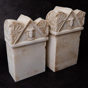 Puede incluir: Dos sujetalibros de piedra color hueso con base rectangular y front&oacute;n triangular. Cada front&oacute;n muestra un rostro humano esculpido en relieve en el centro, flanqueado por intrincadas tallas florales con un acabado desgastado.