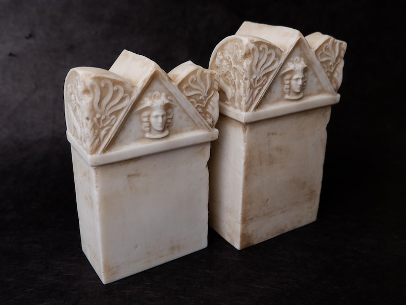 Puede incluir: Dos sujetalibros de piedra color hueso con base rectangular y front&oacute;n triangular. Cada front&oacute;n muestra un rostro humano esculpido en relieve en el centro, flanqueado por intrincadas tallas florales con un acabado desgastado.
