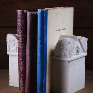 Puede incluir: Un par de sujetalibros de piedra color hueso con detalles en relieve tallados de figuras y follaje, sosteniendo tres libros. Los libros incluyen un volumen rojo oscuro titulado Ruhun &Ouml;l&uuml;m&uuml;, un lomo marr&oacute;n oscuro liso y un lomo azul junto a un libro crema.
