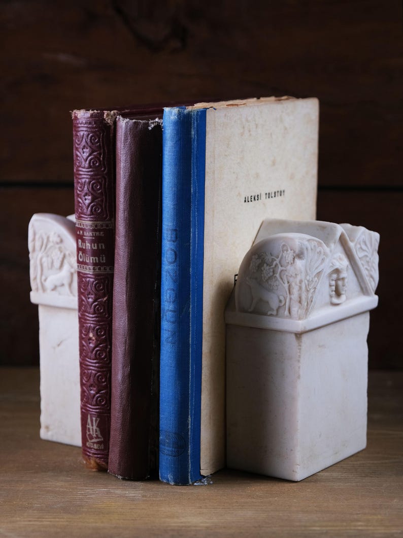 Puede incluir: Un par de sujetalibros de piedra color hueso con detalles en relieve tallados de figuras y follaje, sosteniendo tres libros. Los libros incluyen un volumen rojo oscuro titulado Ruhun &Ouml;l&uuml;m&uuml;, un lomo marr&oacute;n oscuro liso y un lomo azul junto a un libro crema.