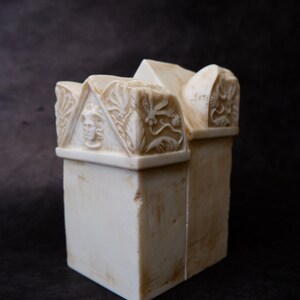 Puede incluir: Una escultura de piedra de color crema con una base rectangular y una secci&oacute;n superior en forma de tejado. El front&oacute;n frontal muestra un rostro humano tallado en relieve, flanqueado por intrincados motivos florales y de hojas.