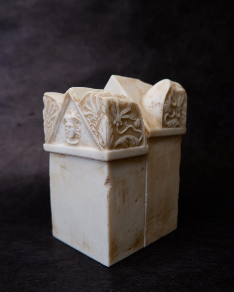 Puede incluir: Una escultura de piedra de color crema con una base rectangular y una secci&oacute;n superior en forma de tejado. El front&oacute;n frontal muestra un rostro humano tallado en relieve, flanqueado por intrincados motivos florales y de hojas.